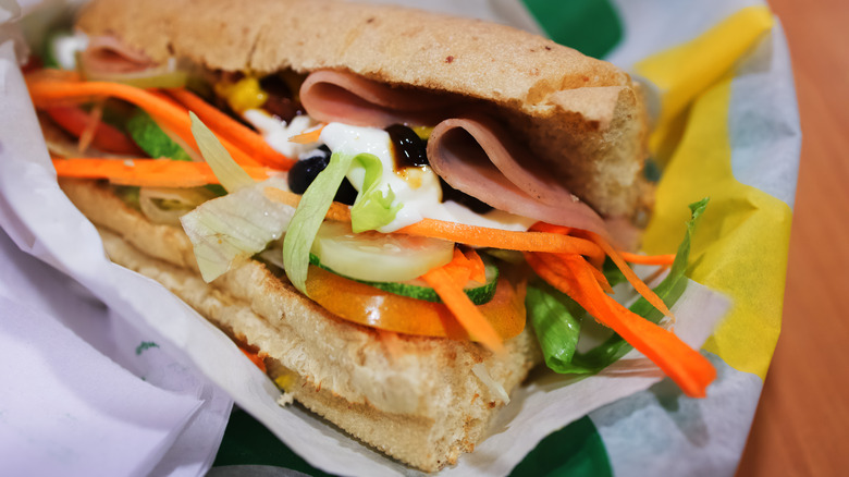 A Subway sandwich filled with tomato, cucumber, olives, carrots, lettuce, and a slice of ham