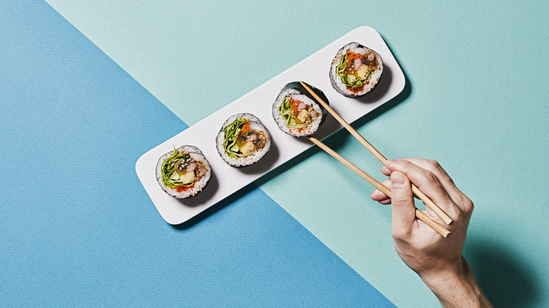 A hand holding chopsticks picks up a piece of sushi from a rectangular serving dish