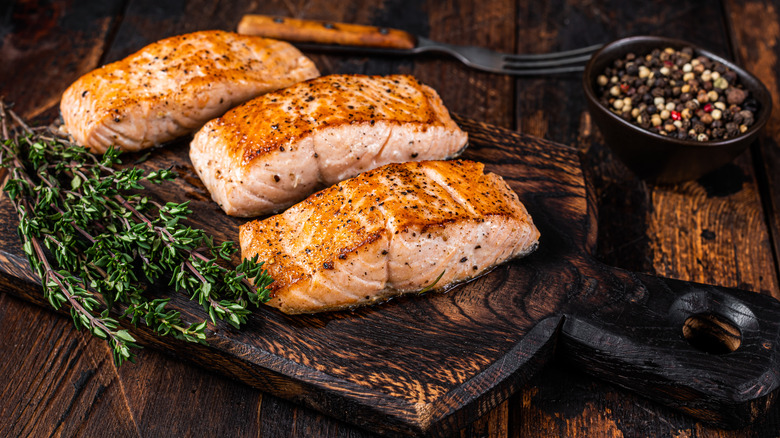 salmon on a wooden cutting board with a sprig of herbs next to a bowl of peppercorns