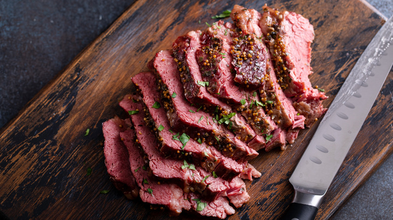 Sliced corned beef with seasoning and garnish on a wooden cutting board with a long knife near. All on a dark table.