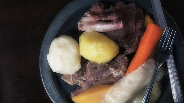 Jiggs Dinner plated on a dark background, with salt beef, potatoes, carrots, and cabbage, with utensils on the plate.