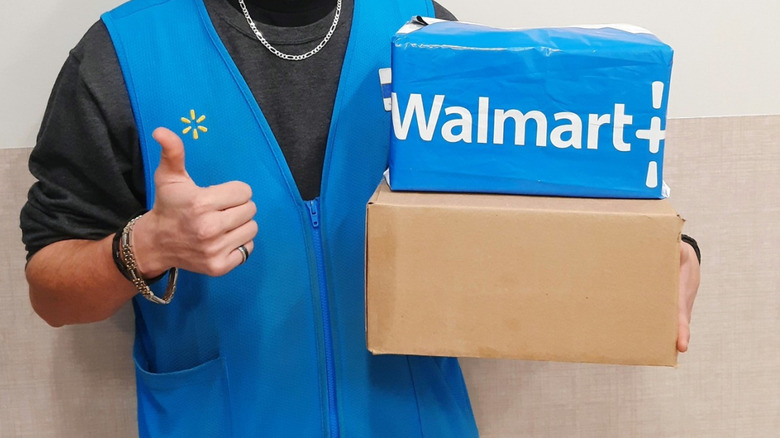 A Walmart employee gives a thumbs up while holding a box with the Walmart+ logo