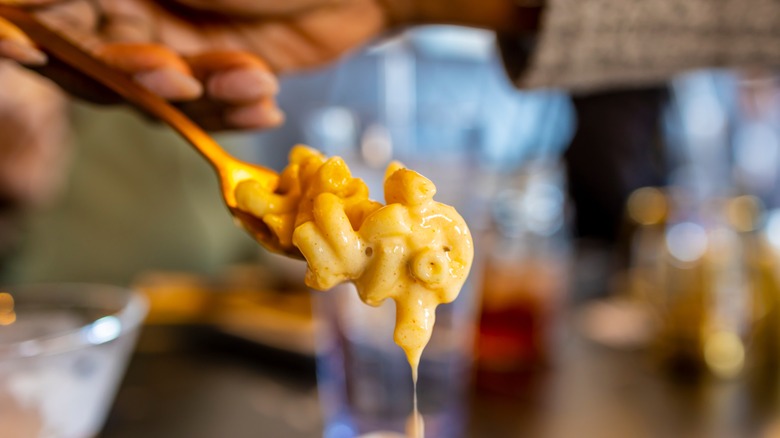 A hand holds a forkful of macaroni and cheese, with sauce dripping from the noodles