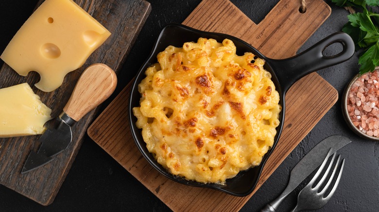 Macaroni and cheese plated in a cast iron skilled sits on a wooden cutting board, with Swiss cheese, utensils, pink Himalayan salt, and herbs sitting adjacent