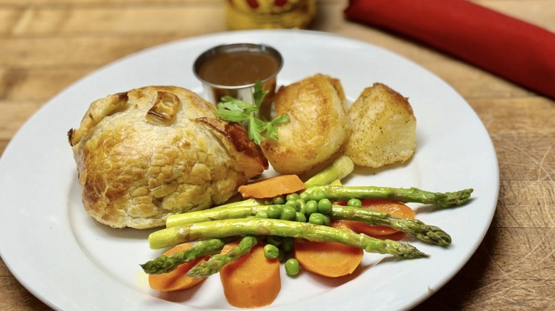 A small, individually made beef Wellington sits on a plate with roasted vegetables and potatoes, with a side of gravy.