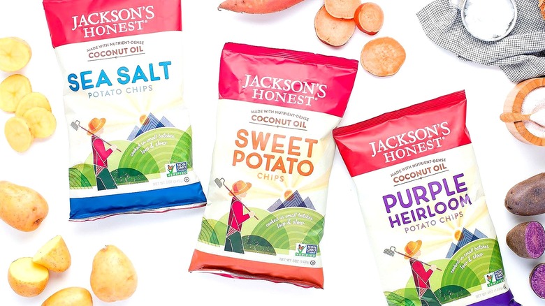 Three bags of Jackson's Honest potato chips surrounded by raw sliced potatoes