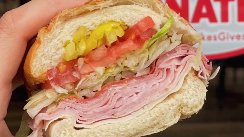 A hand holding up a Jersey Mike's sub with meat, cheese, lettuce, and banana peppers