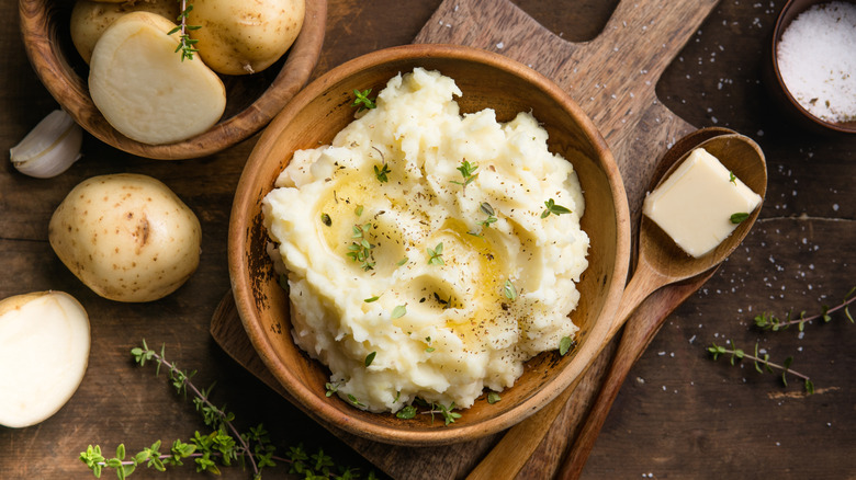 Mashed potatoes in a wooden bowl next to sliced potatoes, a spoonful of butter, and salt.