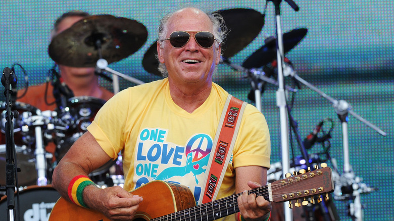 Jimmy Buffett plays guitar in front of a band in an outdoor beach setting.