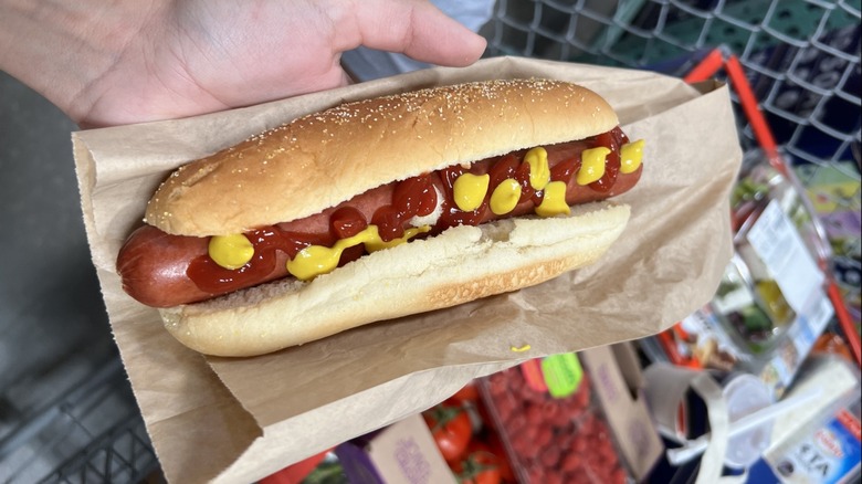 A hand holds a Costco food court hot dog with ketchup and mustard