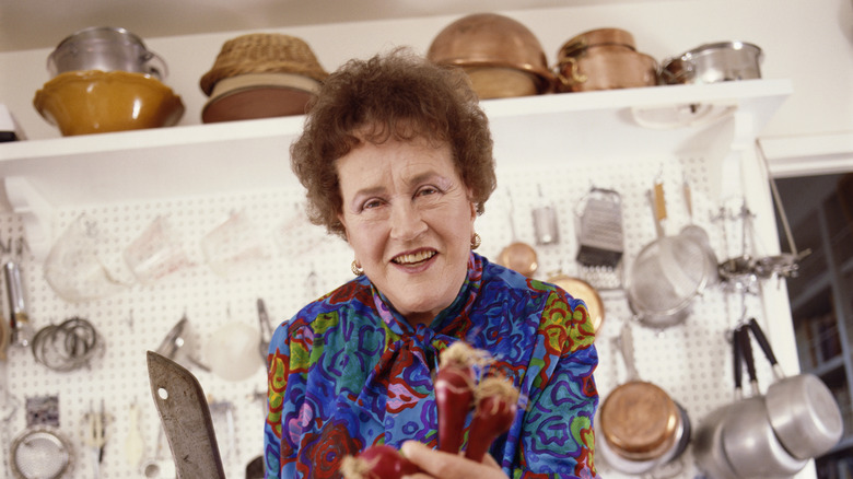 A color photo of Julia Child smiling in her kitchen, with the handles of rolling pins near her and pots and pans on the wall behind her