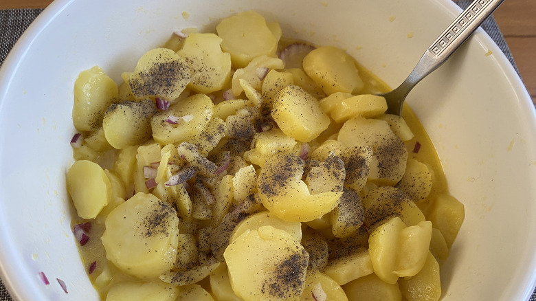 A potato salad is made in a white bowl with a silver spoon; the potatoes are sliced in rounds, with diced red onion, black pepper, and broth also in the bowl