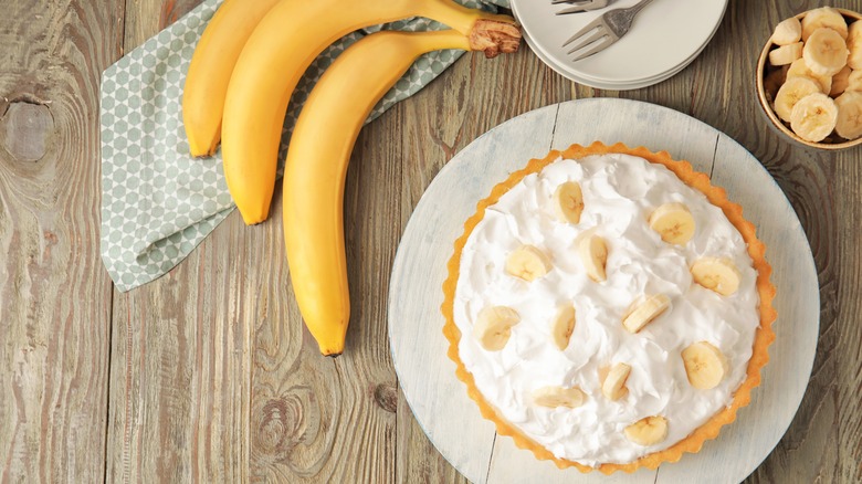 banan cream pie on a light colored plate on a wooden table with whole bananas and sliced bananas