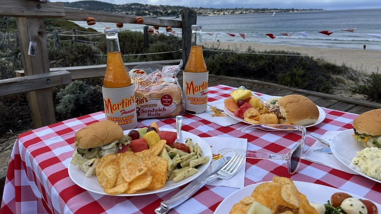 A checkered tablecloth is covered with plated burgers made using Martin's Famous Potato Rolls, with the beach and ocean in the background.
