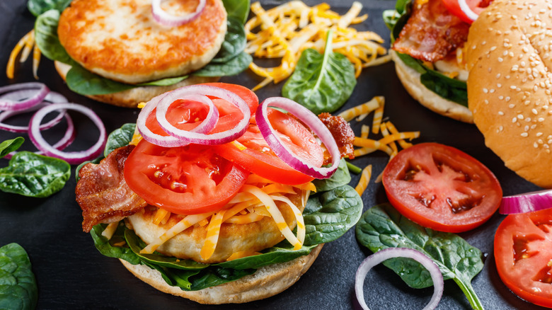 An open face turkey burger, topped with cheese, bacon, tomatoes, fresh red onion, and spinach, surrounded by loose toppings