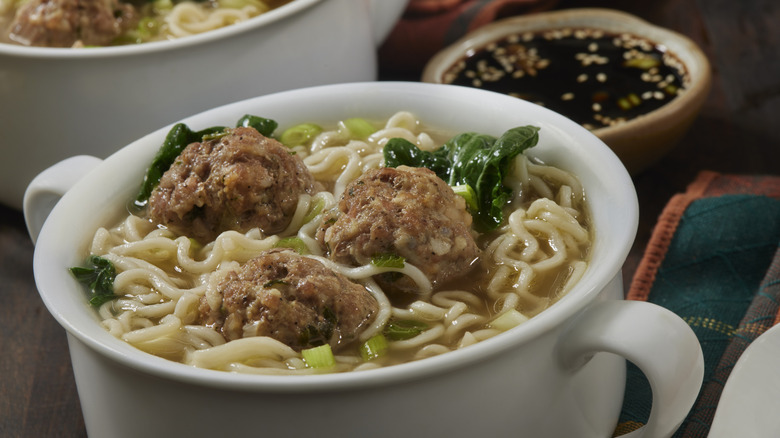 Bowl of pork and shrimp meatball soup with noodles and greens and a bowl of soy sauce behind
