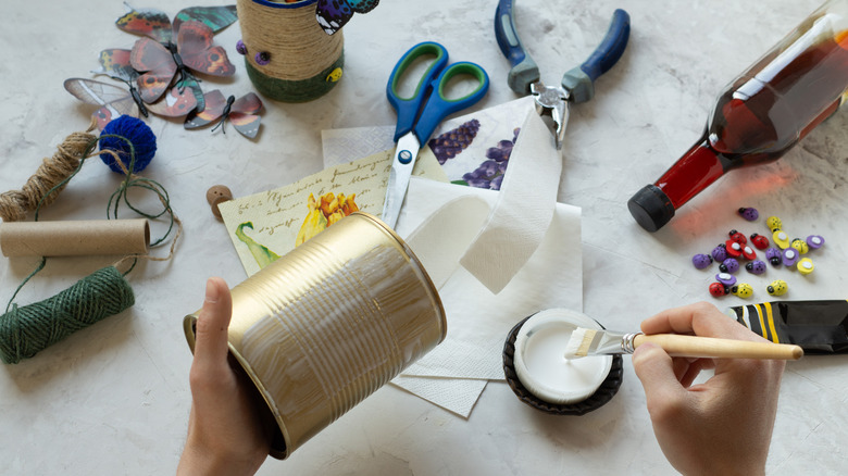 A hand applies glue to an empty soup can, with crafting tools and decorative items sitting around it