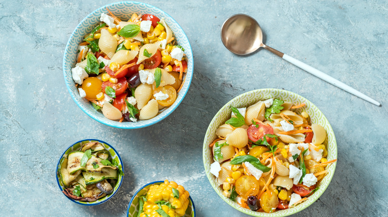 Two bowls of pasta salad on a table with a spoon above and a bowl of cooked zucchini and one of corn on the cob below.