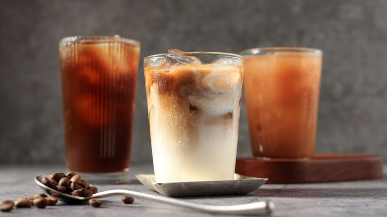 Three kinds of iced coffee sit on a gray counter beside a spoon filled with coffee beans