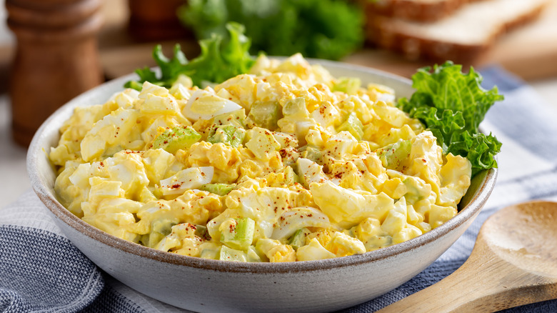 Egg salad in a bowl over lettuce, more lettuce, sliced bread, and pepper grinder in the background.