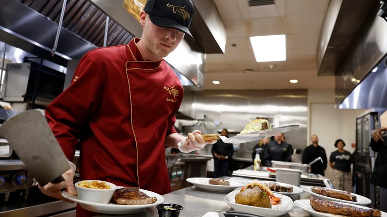 A worker at LongHorn Steakhouse plating several types of food in a commercial kitchen.
