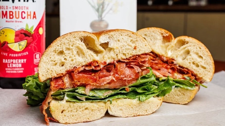 A BLT bagel sits, sliced, on some wax paper next to a bottle of kombucha.