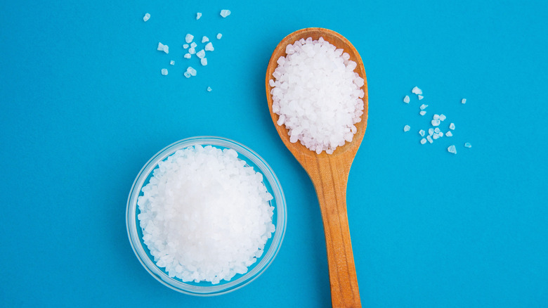 Large grains of salt, some in a glass ramekin, some in a wooden spoon over a blue table