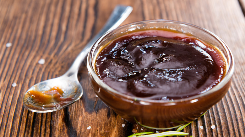 A small bowl of homemade barbecue sauce has a used spoon next to it as well as salt and fresh rosemary