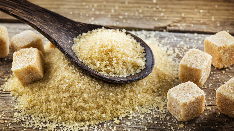 A wooden spoon filled with turbinado sugar sits atop a small mound of turbinado sugar on a rustic wooden surface, with cubes of turbinado sugar surrounding it