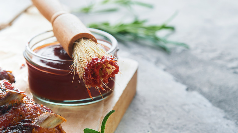 Bowl of barbecue sauce on table with a basting brush sit next to fresh herbs and barbecued ribs