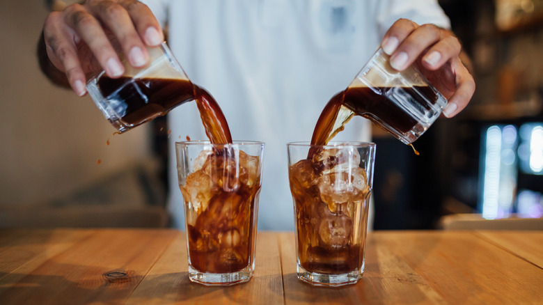 A person pours coffee over ice in clear glass cups