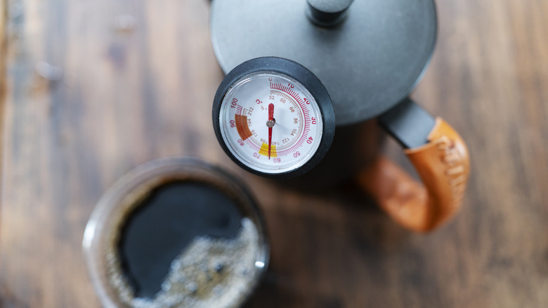 A coffee pot with thermometer sits beside a cup of black coffee