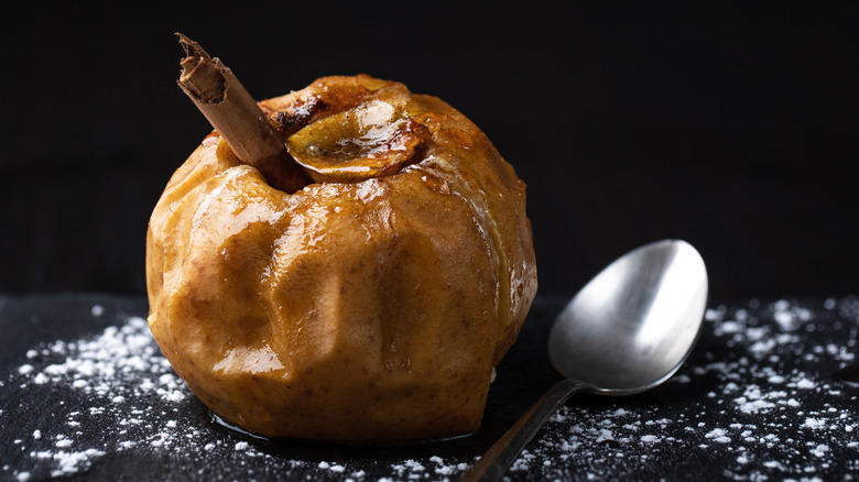 A baked apple with a cinnamon stick on a black table dusted with powdered sugar