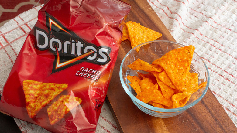 Open bag of Doritos beside a glass bowl of nacho cheese chips on a wooden plank