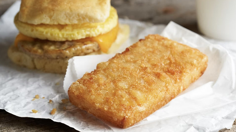 Hash brown served on paper in front of a sausage, egg, and cheese sandwich