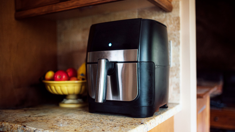 An air fryer sits on a counter near a bowl of fruit