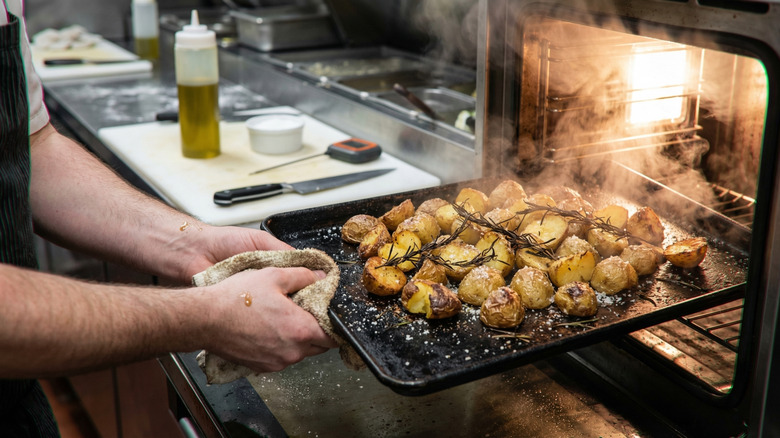 A person pulls a tray of roast potatoes with burnt rosemary from a smoking oven