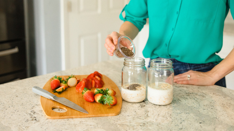 A woman adds chia seeds to her overnight oats next to a cutting board with fresh strawberries