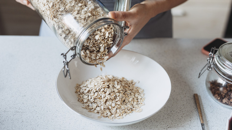 A person pours rolled oats from a glass container into a white bowl