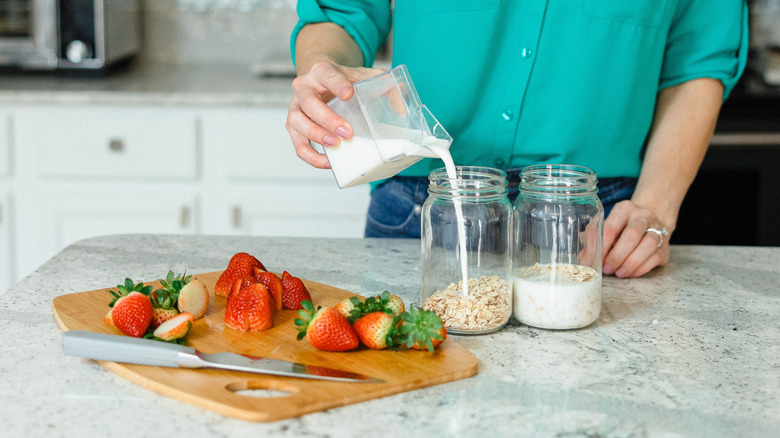 A woman pours milk into a jar of oats next to a cutting board with strawberries