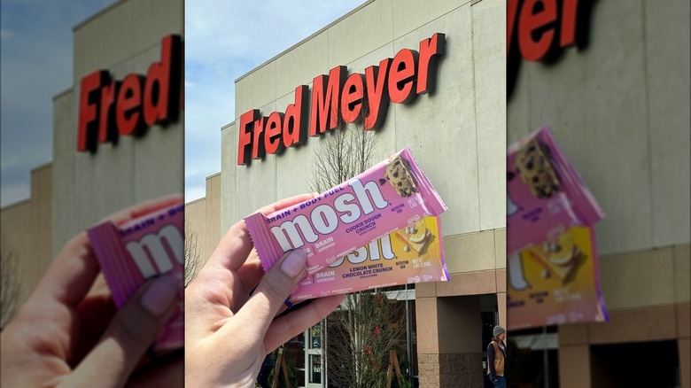 A hand is shown holding two Mosh protein bars in front of a Fred Meyer storefront