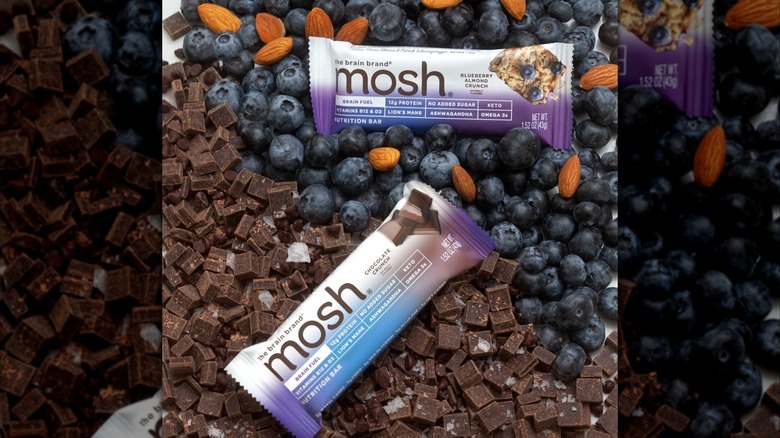 A Mosh Blueberry Almond Crunch bar and a Mosh Chocolate Crunch bar lay atop blueberries, almonds, and chocolate chunks