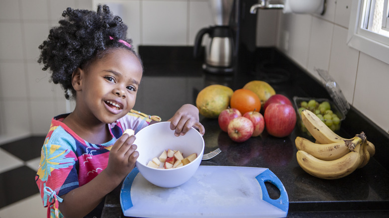 A little girl with a side ponytail eats slices of apple out of a bowl next to apples, oranges, bananas, and grapes