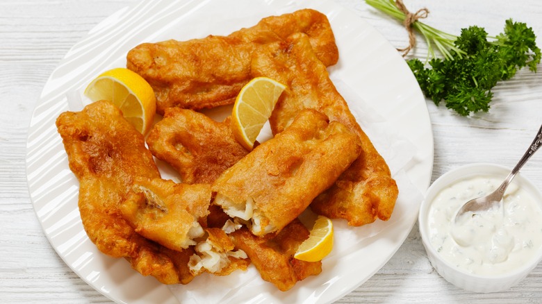 A platter of beer-battered fish sits on a white plate next to a dish of tartare sauce and a sprig of herbs