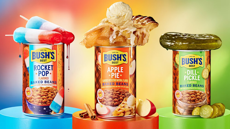 Cans of Bush's Rocket Pop, Apple Pie, and Dill Pickle baked beans sit on colored pedestals, with two Rocket Pops, a wedge of apple pie ala mode, and a dill pickle atop the cans.