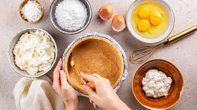 Cheesecake crust being formed by hands, surrounded by cheesecake ingredients