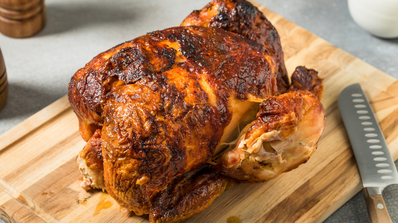 A rotisserie chicken sits on a wooden cutting board next to a knife