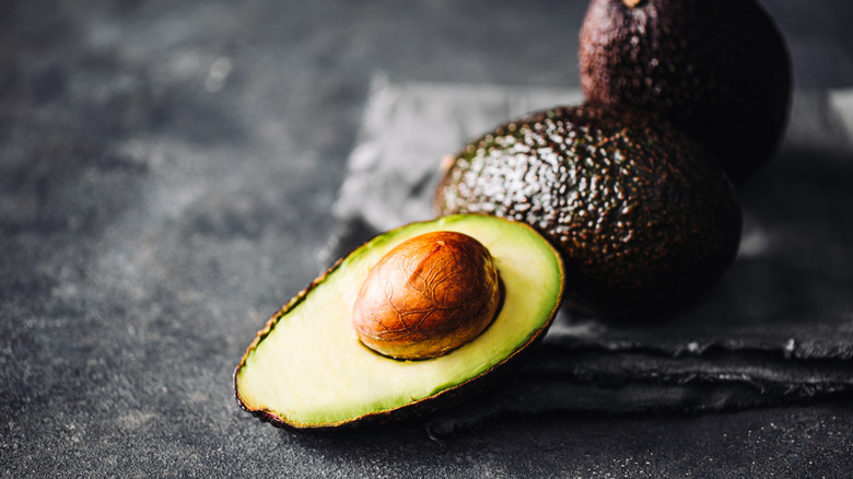 A halved avocado with the pit, with two whole avocados in the background