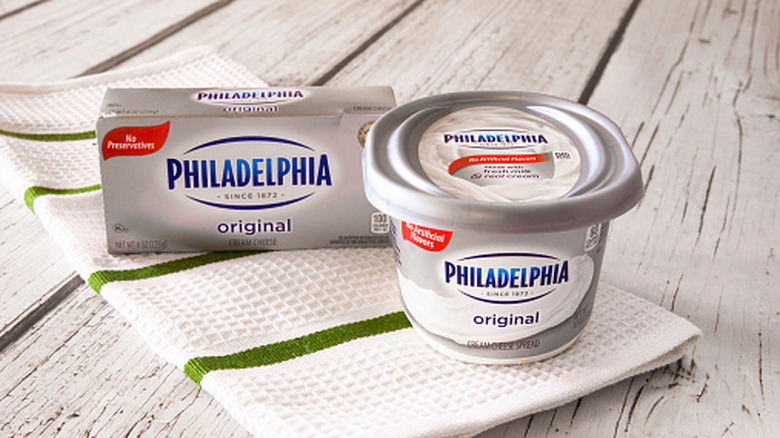 A small container of cream cheese sits next to a block of cream cheese.