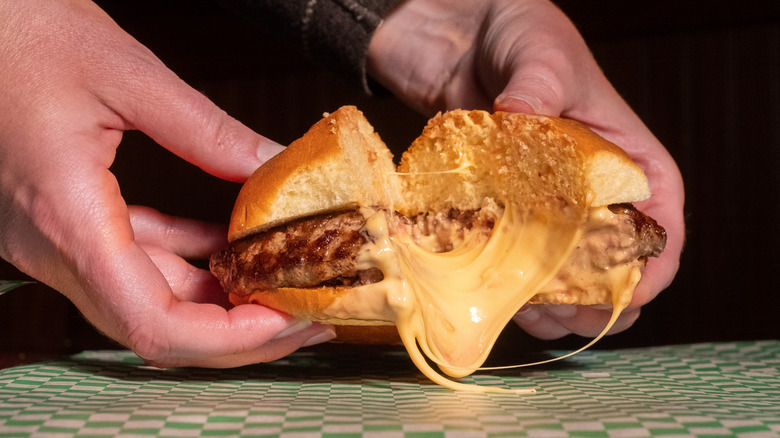 Gooey, melted cheese oozes from between the halves of a stuffed burger, held up by two hands.